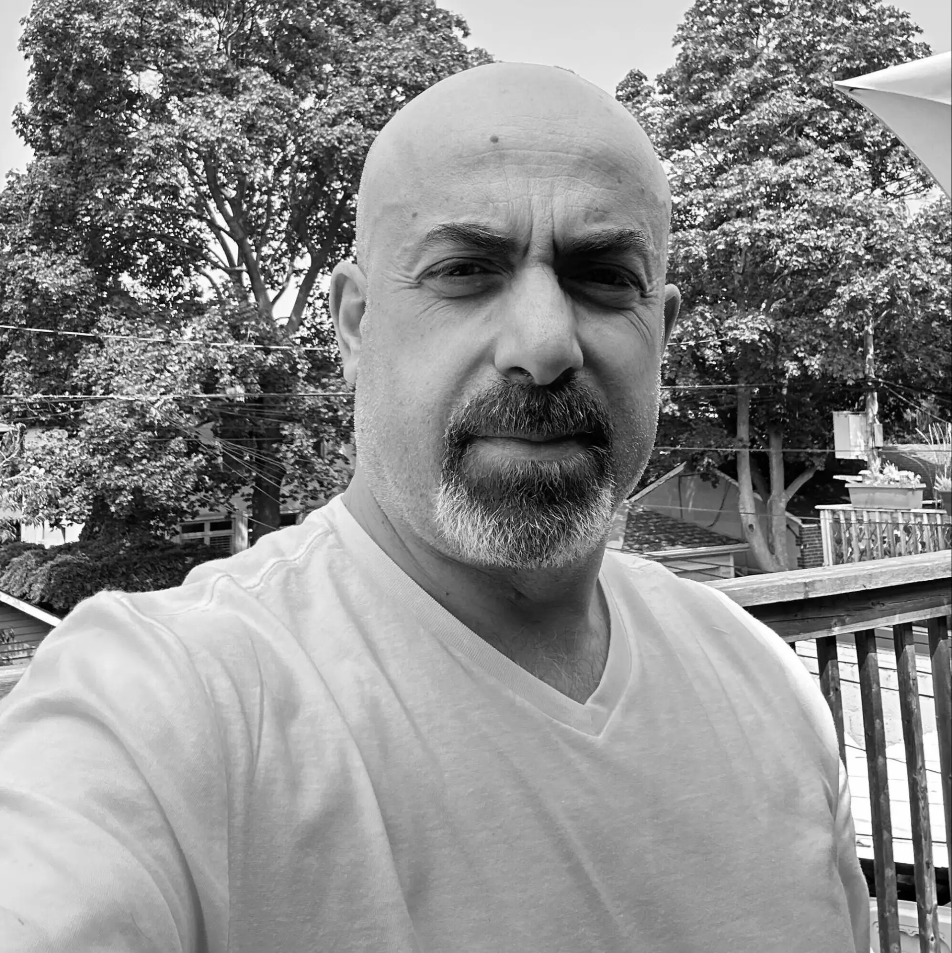 a man with a bald head and a goatee wearing a white t shirt, standing outdoors on a wooden deck. in the background, there are trees and a blue sky. at floka hair salon on bayview in toronto